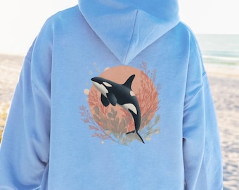 May include: Light blue hoodie featuring a graphic of a black and white orca whale swimming in front of a coral reef design. The orca is set against a peach-colored circle. The hoodie is made of a soft material.