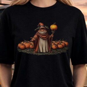 May include: A black t-shirt featuring a graphic illustration of a frog dressed as a wizard in a brown robe and pointed hat. The frog holds a staff topped with a pumpkin, standing amidst a patch of orange pumpkins on a dark, textured ground.