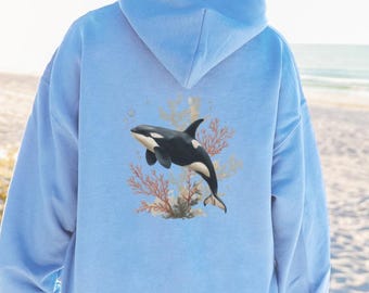 May include: A light blue hooded sweatshirt featuring a graphic print on the back. The design depicts a black and white orca swimming amidst coral reef elements in shades of orange and tan. The hoodie has long sleeves and a large hood.