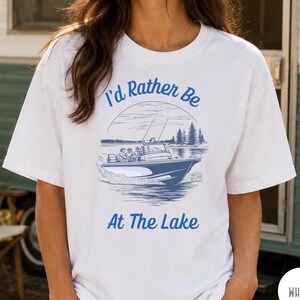 Op de afbeelding: Wit T-shirt met een blauwe afbeelding van een boot op een meer, met de tekst "I'd Rather Be At The Lake" in het blauw. Het shirt is een casual stijl, geschikt voor buitenactiviteiten.
