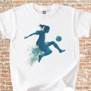 May include: White t-shirt with a blue silhouette of a soccer player kicking a ball. The design has a watercolour effect, with blue and white splashes. The shirt has a crew neck and short sleeves. The word "WHITE" is printed on the bottom right.