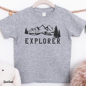 May include: A grey t-shirt featuring a black mountain and tree graphic with the word "EXPLORER". The shirt is displayed on a wooden hanger. A white soft toy and a pink fluffy item are in the background.