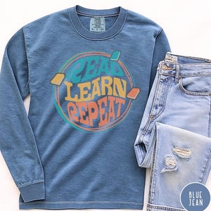 May include: A blue long-sleeve shirt with the words "READ LEARN REPEAT" in a colorful circular design. The shirt is paired with light blue denim jeans with distressed details. A small blue circle with the words "BLUE JEAN" is visible.