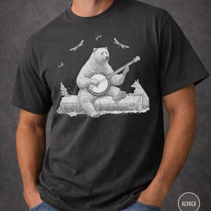 May include: Dark gray t-shirt featuring a whimsical illustration of a bear playing a banjo while seated on a log, with a fox nearby. Dragonflies are in the background. The design is in black and white.