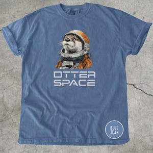 K&ouml;nnte beinhalten: Blaues T-Shirt mit dem wei&szlig;en Schriftzug "OTTER SPACE". Das Shirt zeigt eine Illustration eines Otters mit orangefarbenem Weltraumhelm und -anzug. Ein kleiner blauer Kreis mit "BLUE JEAN" befindet sich unten rechts. Baumwollkleidung.