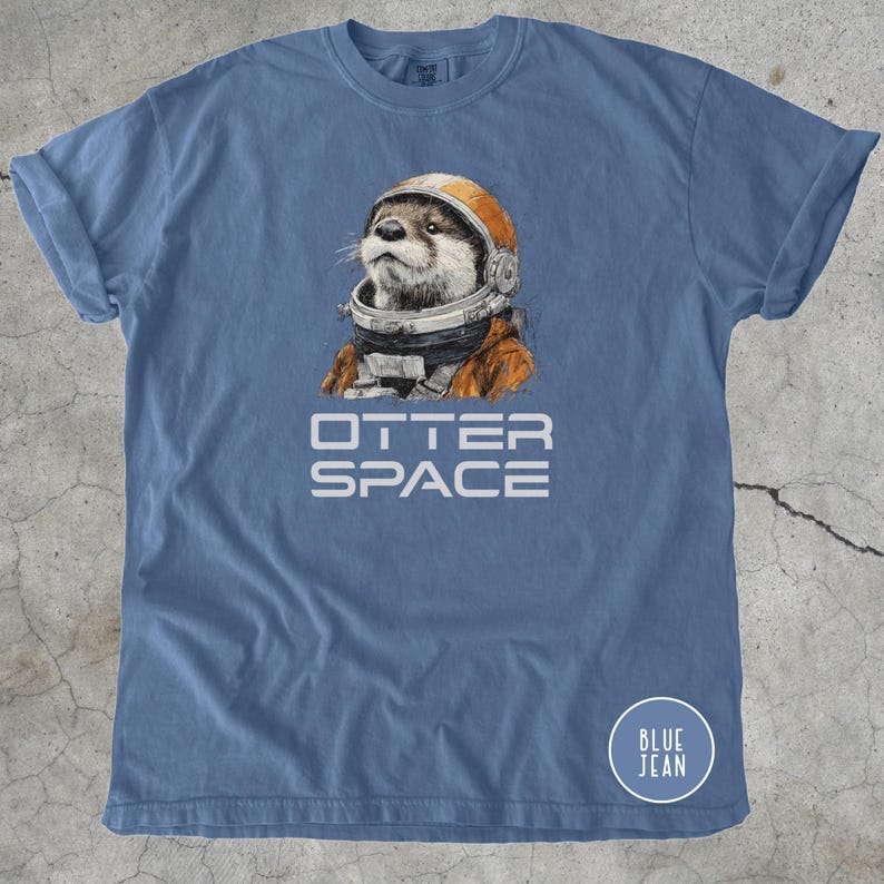 K&ouml;nnte beinhalten: Blaues T-Shirt mit dem wei&szlig;en Schriftzug "OTTER SPACE". Das Shirt zeigt eine Illustration eines Otters mit orangefarbenem Weltraumhelm und -anzug. Ein kleiner blauer Kreis mit "BLUE JEAN" befindet sich unten rechts. Baumwollkleidung.