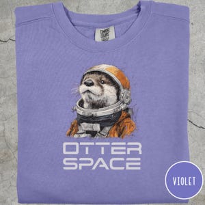 May include: Violet crewneck sweatshirt featuring a graphic of an otter wearing an astronaut helmet and suit. The text "OTTER SPACE" is printed below the otter. The sweatshirt has a round neckline and long sleeves.