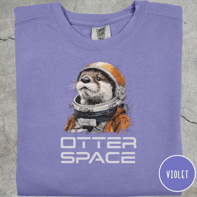 May include: Violet crewneck sweatshirt featuring a graphic of an otter wearing an astronaut helmet and suit. The text "OTTER SPACE" is printed below the otter. The sweatshirt has a round neckline and long sleeves.
