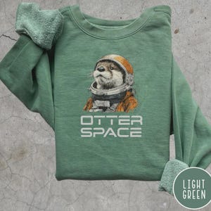 K&ouml;nnte beinhalten: Hellgr&uuml;nes Sweatshirt mit einem Aufdruck eines Otters mit Astronautenhelm. Darunter steht der Text "OTTER SPACE". Das Sweatshirt hat einen Rundhalsausschnitt und lange &Auml;rmel. Die &Auml;rmel sind hochgekrempelt.