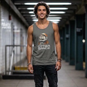 K&ouml;nnte beinhalten: Graues Tanktop mit einem Otter in einem Weltraumanzug-Grafik und dem Text "OTTER SPACE". Das &auml;rmellose Shirt ist aus einem weichen Material gefertigt und wird mit schwarzen Jeans kombiniert. Die Person steht in einer U-Bahn-Station.