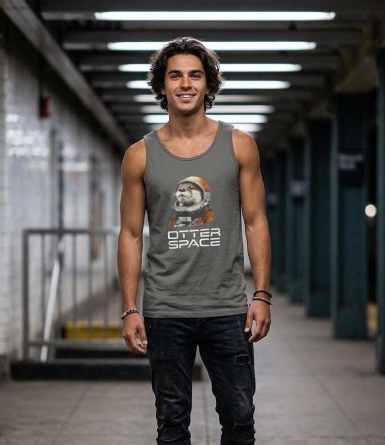 K&ouml;nnte beinhalten: Graues Tanktop mit einem Otter in einem Weltraumanzug-Grafik und dem Text "OTTER SPACE". Das &auml;rmellose Shirt ist aus einem weichen Material gefertigt und wird mit schwarzen Jeans kombiniert. Die Person steht in einer U-Bahn-Station.