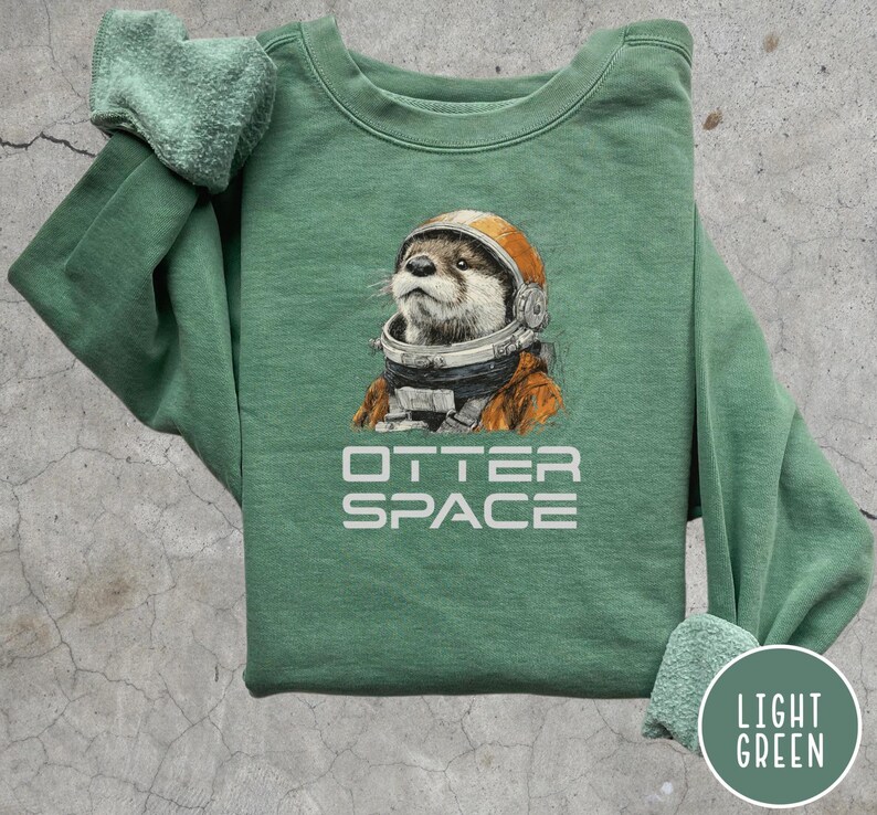 May include: Light green sweatshirt featuring an otter wearing an astronaut helmet and suit. The text "OTTER SPACE" is printed in white below the otter illustration. The sweatshirt has a crew neck and long sleeves.