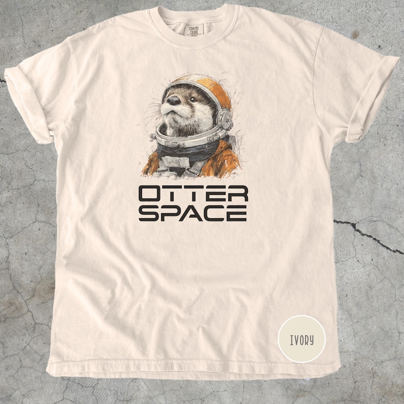 K&ouml;nnte beinhalten: Elfenbeinfarbenes T-Shirt mit einer detaillierten Illustration eines Otters mit Weltraumhelm und -anzug. Der Text "OTTER SPACE" ist in fetten, schwarzen Buchstaben unter dem Bild aufgedruckt. Das Shirt ist aus weichem Material.
