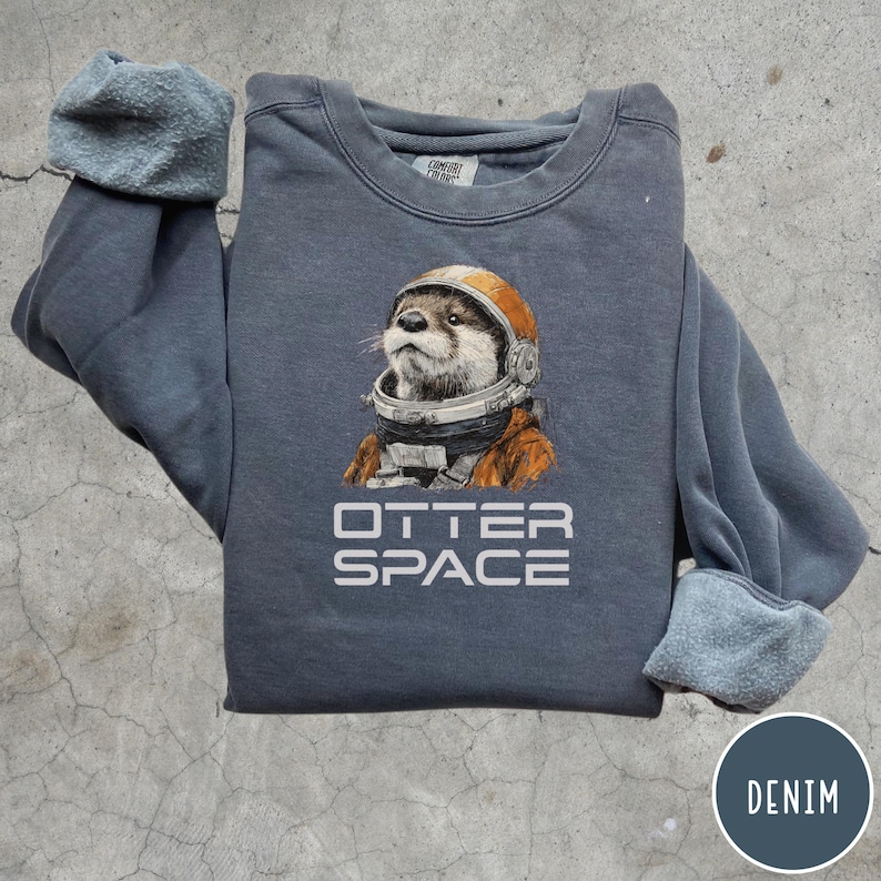May include: A denim blue sweatshirt featuring a graphic of an otter wearing an astronaut helmet. The text "OTTER SPACE" is printed below the otter. The sweatshirt has a crew neck and long sleeves with ribbed cuffs.