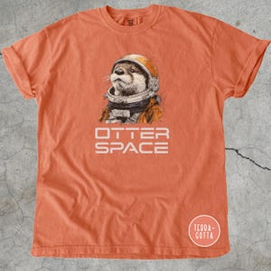K&ouml;nnte beinhalten: Orangefarbenes T-Shirt mit einer detaillierten Illustration eines Otters mit Weltraumhelm und -anzug. Der Text "OTTER SPACE" ist darunter gedruckt. Das T-Shirt hat hochgekrempelte &Auml;rmel und einen Rundhalsausschnitt.