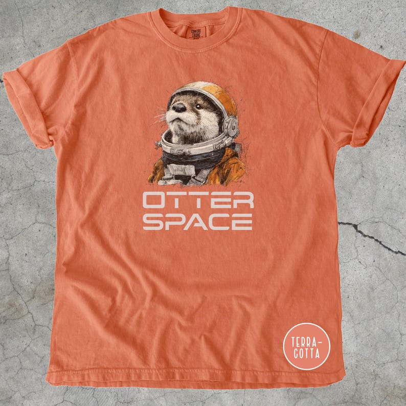 K&ouml;nnte beinhalten: Orangefarbenes T-Shirt mit einer detaillierten Illustration eines Otters mit Weltraumhelm und -anzug. Der Text "OTTER SPACE" ist darunter gedruckt. Das T-Shirt hat hochgekrempelte &Auml;rmel und einen Rundhalsausschnitt.