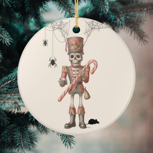 May include: A circular Christmas ornament with a skeleton toy soldier design. The soldier is in a red and blue uniform and holds a candy cane. The ornament has a cream background with black spiders, cobwebs, and a mouse. A gold string is at the top.