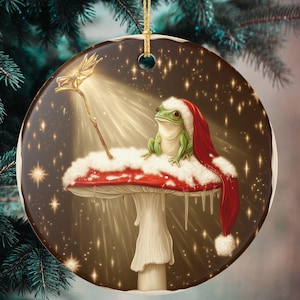 Pode incluir: Enfeite de Natal redondo com um sapo verde usando um gorro de Papai Noel, sentado em um cogumelo vermelho e branco. Uma varinha dourada emite luz. O fundo é marrom escuro com estrelas brilhantes e um fio dourado para pendurar.