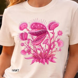Puede incluir: Camiseta de color marfil con una vibrante ilustración rosa de una Venus atrapamoscas. El diseño muestra una anatomía detallada de la planta con trampas abiertas y delicados capullos florales. La palabra "IVORY" está impresa en la parte inferior. Una prenda de vestir de temática botánica.