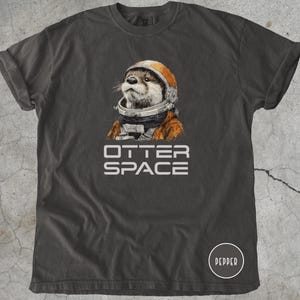 K&ouml;nnte beinhalten: Dunkelgraues T-Shirt mit einer Illustration eines Otters, der einen Astronautenhelm und -anzug tr&auml;gt. Der Text "OTTER SPACE" ist darunter in Wei&szlig; gedruckt. Das T-Shirt hat hochgekrempelte &Auml;rmel und einen Rundhalsausschnitt.