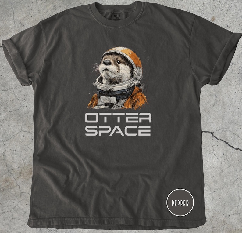 K&ouml;nnte beinhalten: Dunkelgraues T-Shirt mit einer Illustration eines Otters, der einen Astronautenhelm und -anzug tr&auml;gt. Der Text "OTTER SPACE" ist darunter in Wei&szlig; gedruckt. Das T-Shirt hat hochgekrempelte &Auml;rmel und einen Rundhalsausschnitt.