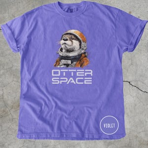 K&ouml;nnte beinhalten: Violettes T-Shirt mit detaillierter Illustration eines Otters mit Astronautenhelm und -anzug. Der Text "OTTER SPACE" ist darunter gedruckt. Ein Kreis mit dem Wort "VIOLET" befindet sich in der unteren rechten Ecke.