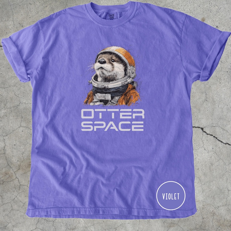 K&ouml;nnte beinhalten: Violettes T-Shirt mit detaillierter Illustration eines Otters mit Astronautenhelm und -anzug. Der Text "OTTER SPACE" ist darunter gedruckt. Ein Kreis mit dem Wort "VIOLET" befindet sich in der unteren rechten Ecke.