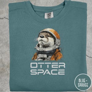 May include: Blue-spruce colored sweatshirt with a graphic of an otter wearing an astronaut helmet and suit. The text "OTTER SPACE" is printed below the otter. The sweatshirt has a crew neck and long sleeves.