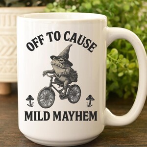 May include: A white ceramic mug featuring a black ink illustration of a frog wearing a pointed hat and riding a bicycle. The text above the frog reads "OFF TO CAUSE" and the text below reads "MILD MAYHEM," with small mushroom icons on each side.