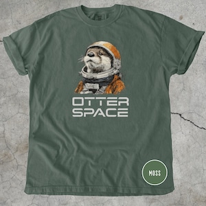 K&ouml;nnte beinhalten: Ein moosgr&uuml;nes T-Shirt mit einer detaillierten Illustration eines Otters mit Astronautenhelm und -anzug. Der Text "OTTER SPACE" ist wei&szlig; unter dem Otter aufgedruckt. Ein kleiner Kreis mit dem Wort "MOSS" befindet sich in der unteren rechten Ecke.