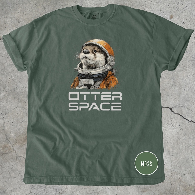 K&ouml;nnte beinhalten: Ein moosgr&uuml;nes T-Shirt mit einer detaillierten Illustration eines Otters mit Astronautenhelm und -anzug. Der Text "OTTER SPACE" ist wei&szlig; unter dem Otter aufgedruckt. Ein kleiner Kreis mit dem Wort "MOSS" befindet sich in der unteren rechten Ecke.