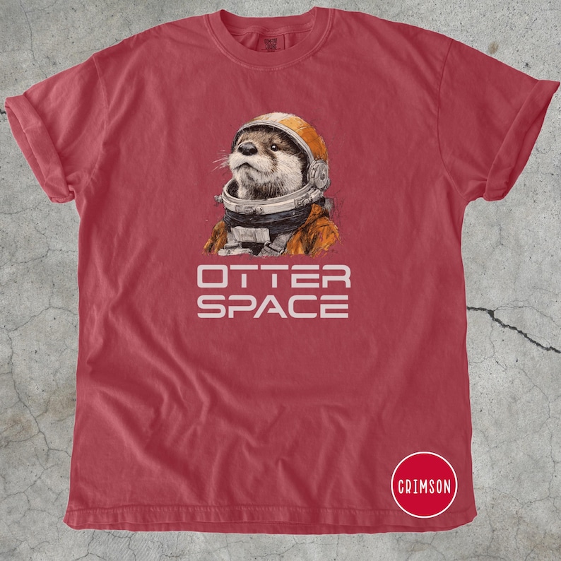 K&ouml;nnte beinhalten: Rotes T-Shirt mit einer Illustration eines Otters, der einen Astronautenhelm und -anzug tr&auml;gt. Der Text "OTTER SPACE" ist darunter gedruckt. Das T-Shirt hat hochgekrempelte &Auml;rmel und einen Rundhalsausschnitt.