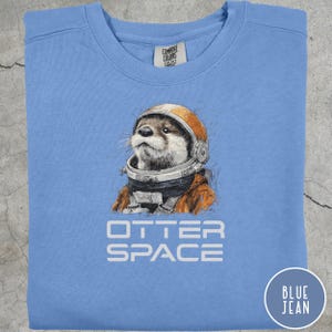 May include: Light blue sweatshirt with a graphic of an otter wearing an astronaut helmet and suit. The text "OTTER SPACE" is printed below the otter. A circular "BLUE JEAN" label is in the bottom right corner.
