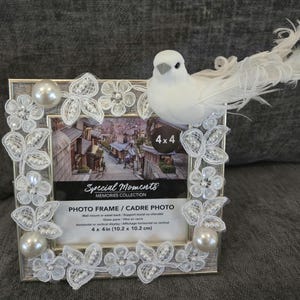Op de afbeelding: Een zilveren fotolijst met witte kant en parelversieringen, met een foto van 10,2 x 10,2 cm. Een witte vogel met een gevederde staart zit bovenop het frame. De lijst is gemarkeerd met "Special Moments" en "Photo Frame".