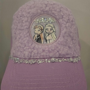 May include: A lavender baseball cap with a fuzzy top and a smooth, curved brim. The cap features a circular image of two cartoon characters and is embellished with rhinestones along the brim.