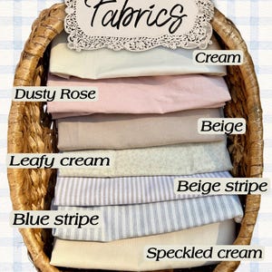 May include: A woven wicker basket containing folded fabric swatches labeled Cream, Dusty Rose, Beige, Leafy cream, Beige stripe, Blue stripe, and Speckled cream. A decorative sign at the top displays the word Fabrics in a script font.