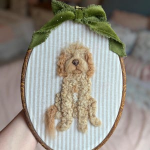 May include: An oval embroidery hoop containing a needle-felted light brown curly-haired dog on a beige and white striped fabric background. The hoop is framed in wood and decorated with a green velvet ribbon tied at the top center.
