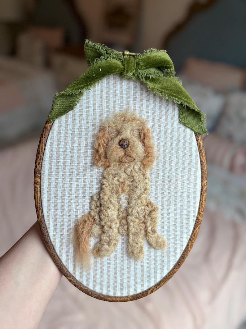 May include: An oval embroidery hoop containing a needle-felted light brown curly-haired dog on a beige and white striped fabric background. The hoop is framed in wood and decorated with a green velvet ribbon tied at the top center.