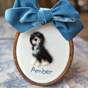 May include: An oval wooden embroidery hoop containing a needle-felted Bernese Mountain Dog puppy on white fabric. The name Amber is embroidered in blue thread below the dog. A large, blue velvet bow is attached to the top of the wooden frame.