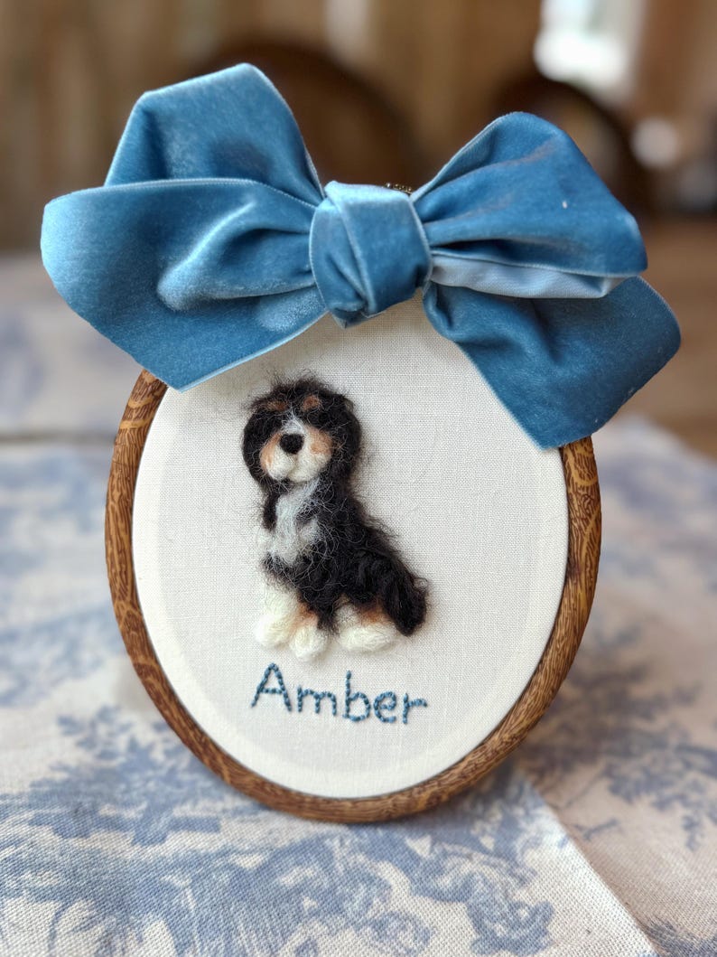 May include: An oval wooden embroidery hoop containing a needle-felted Bernese Mountain Dog puppy on white fabric. The name Amber is embroidered in blue thread below the dog. A large, blue velvet bow is attached to the top of the wooden frame.