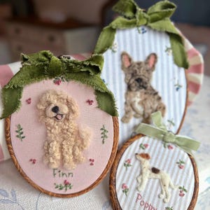 May include: Three oval wooden embroidery hoops featuring needle-felted dog portraits. One hoop shows a cream-colored dog named Finn on pink fabric, another a brindle French Bulldog on blue stripes, and the third a white dog named Poppy on stripes.