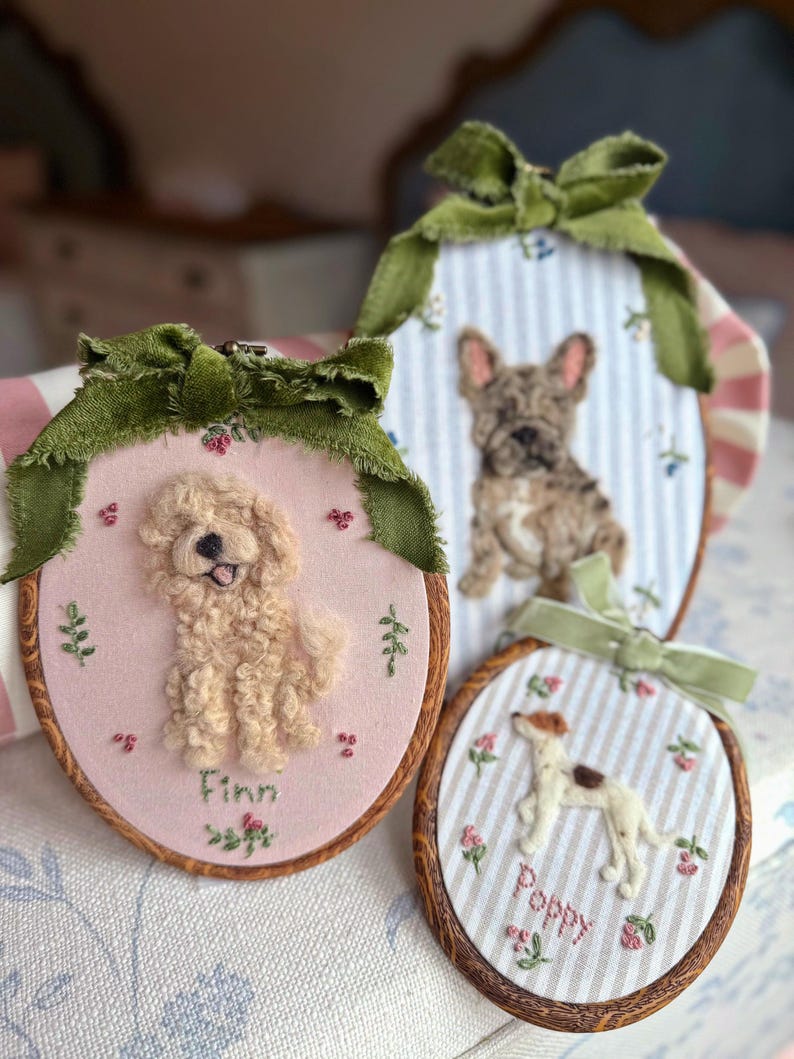 May include: Three oval wooden embroidery hoops featuring needle-felted dog portraits. One hoop shows a cream-colored dog named Finn on pink fabric, another a brindle French Bulldog on blue stripes, and the third a white dog named Poppy on stripes.