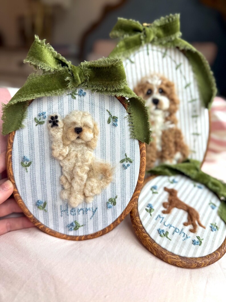 May include: Three oval embroidery hoops featuring needle-felted dog portraits on blue and white striped fabric. Each hoop is topped with a green velvet ribbon and decorated with embroidered blue flowers. The names Henry and Murphy are stitched below.