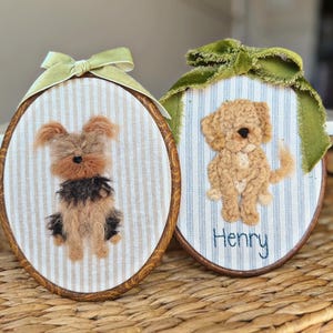 May include: Two oval embroidery hoops featuring needle-felted dog portraits on striped fabric. The left hoop has a light green ribbon and a tan and black dog. The right hoop has a dark green ribbon, a tan dog, and the embroidered name Henry.
