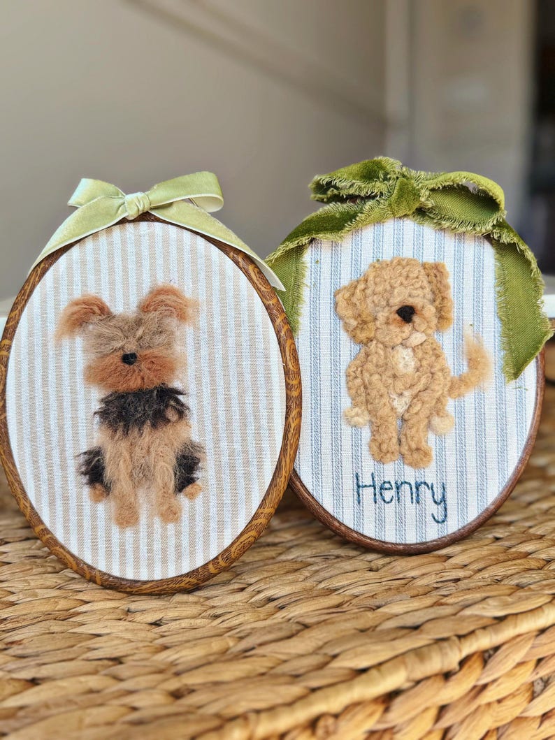 May include: Two oval embroidery hoops featuring needle-felted dog portraits on striped fabric. The left hoop has a light green ribbon and a tan and black dog. The right hoop has a dark green ribbon, a tan dog, and the embroidered name Henry.