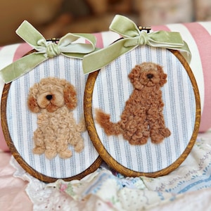 May include: Two oval wooden embroidery hoops featuring needle-felted dogs on blue and white striped fabric. Each hoop is topped with a sage green velvet ribbon bow. One dog is light tan, and the other is a darker reddish-brown, both textured.