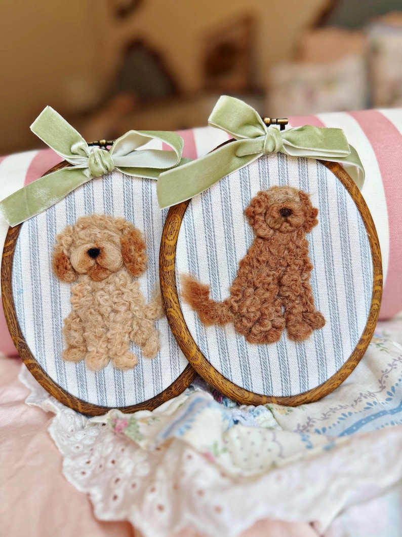 May include: Two oval wooden embroidery hoops featuring needle-felted dogs on blue and white striped fabric. Each hoop is topped with a sage green velvet ribbon bow. One dog is light tan, and the other is a darker reddish-brown, both textured.
