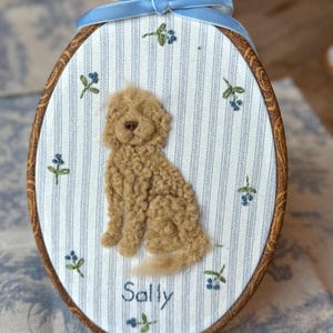 May include: An oval embroidery hoop containing a fabric panel with blue and white vertical stripes. A textured, tan, needle-felted dog sits in the center. Small embroidered blue flowers are scattered around the dog. The name Sally is stitched in blue thread below the dog. A light blue velvet ribbon is tied at the top.