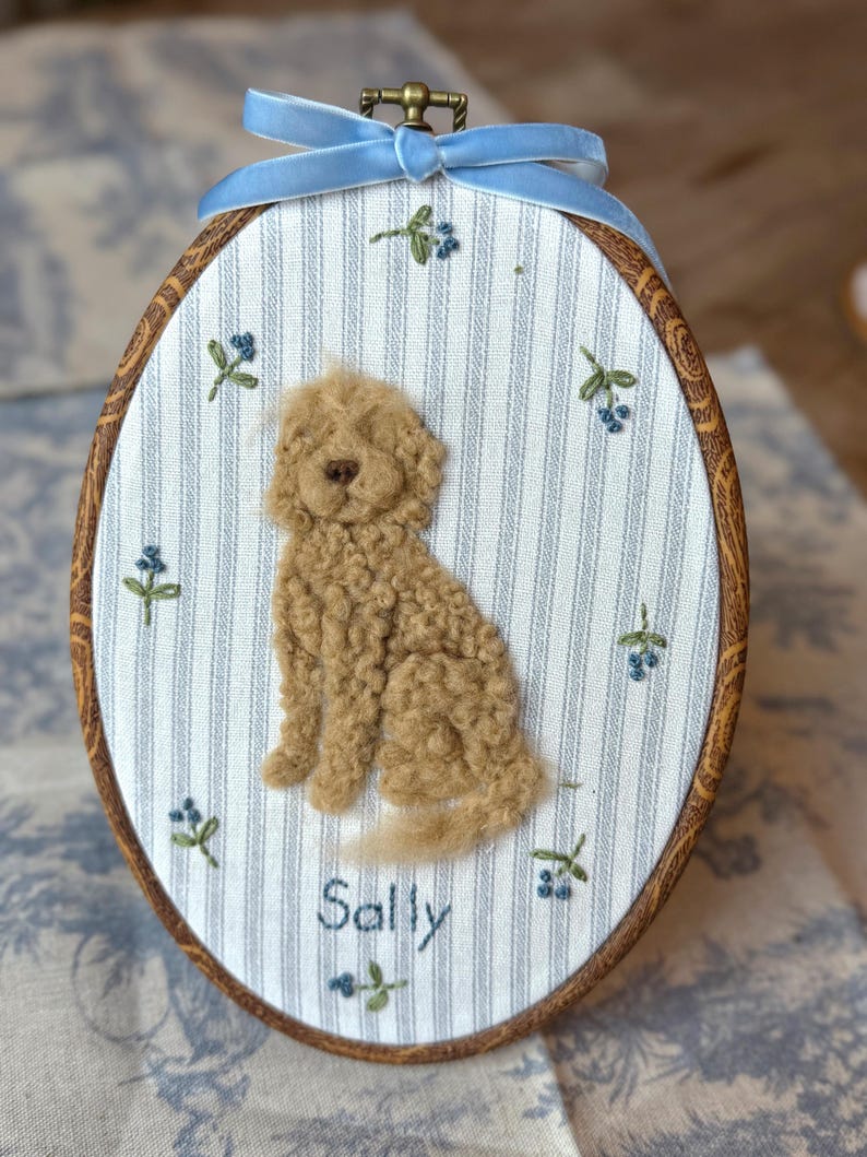 May include: An oval embroidery hoop containing a fabric panel with blue and white vertical stripes. A textured, tan, needle-felted dog sits in the center. Small embroidered blue flowers are scattered around the dog. The name Sally is stitched in blue thread below the dog. A light blue velvet ribbon is tied at the top.