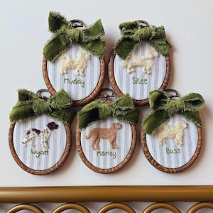 May include: Five oval wooden embroidery hoops featuring needle-felted dog portraits on blue and white striped fabric. Each hoop is topped with a frayed olive green velvet bow and embroidered with the names Muddy, Skee, Wyatt, Marley, and Boss.